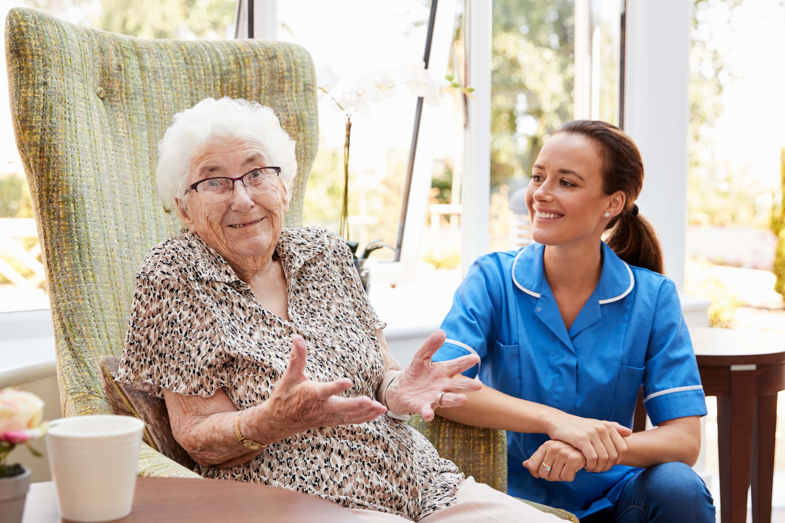 Strange looking elder lady making a gesture at the camera while nurse kneels next to her smiling.