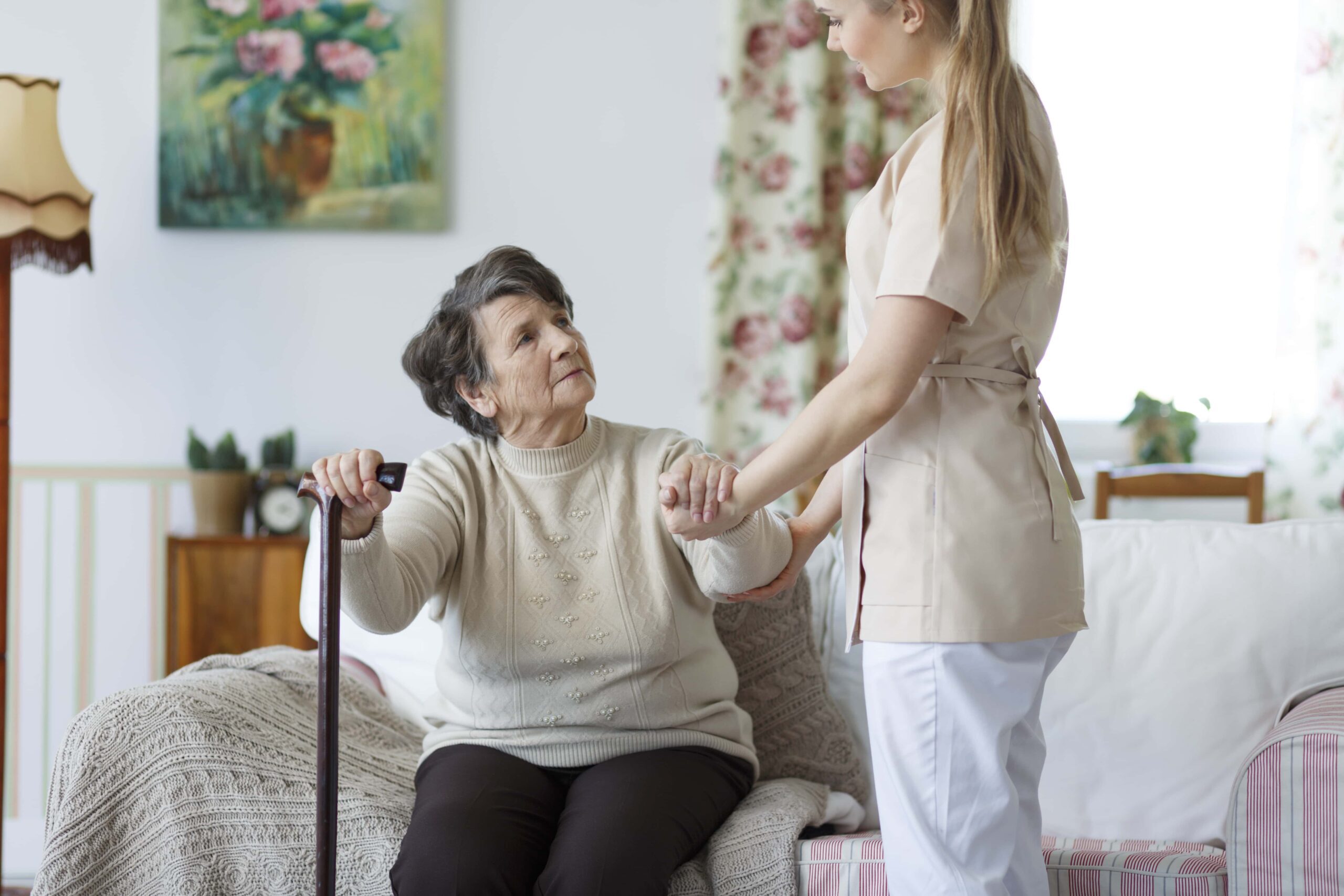 Seated elderly women with cane receiving assistance on standing up from couch.