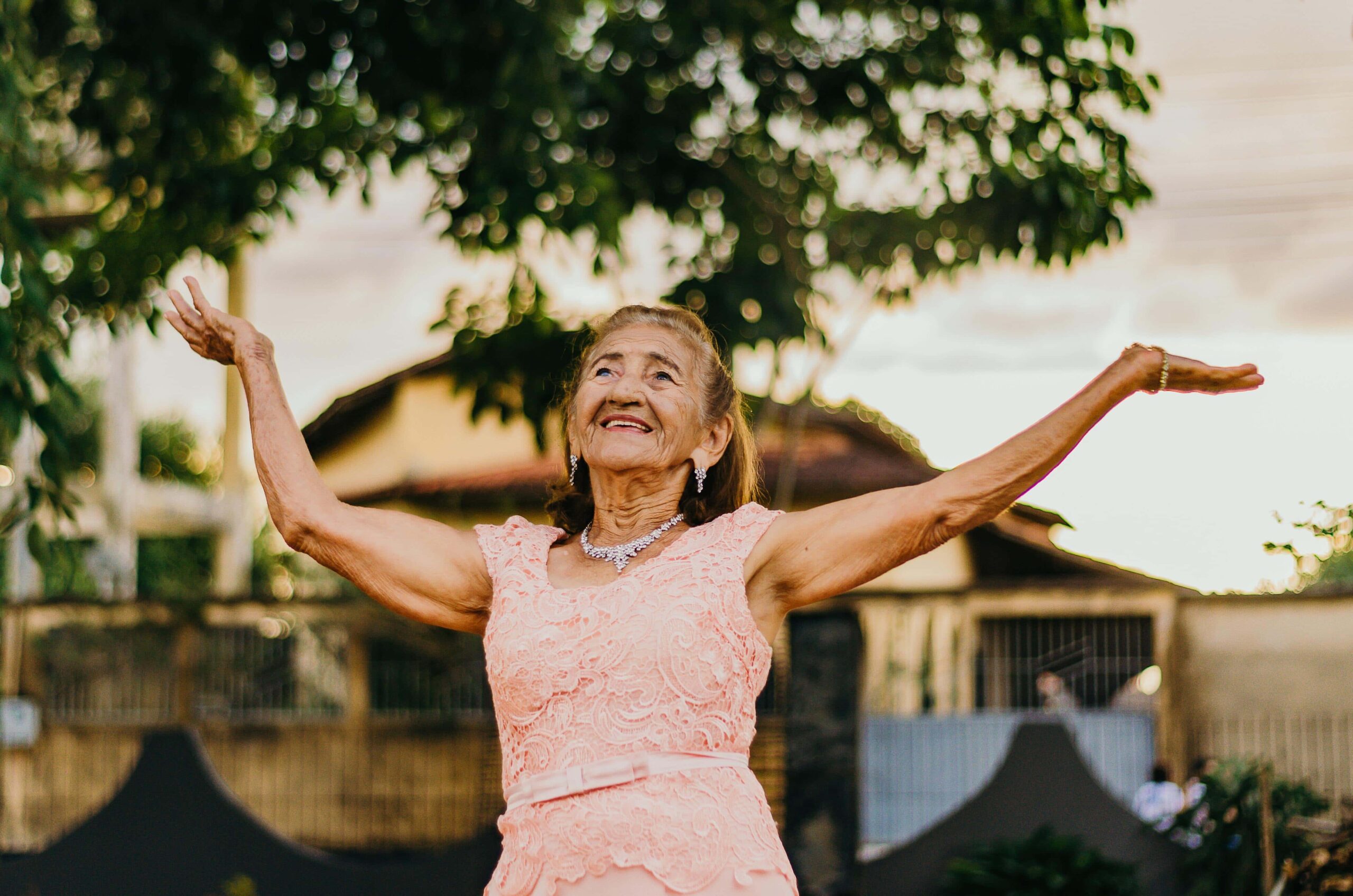 Elderly lady outside with her arms raised to the sky.