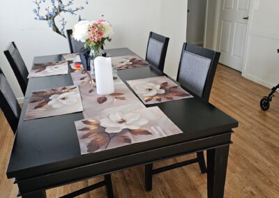 A black dining table with six chairs, floral placemats, a vase of flowers, and a paper towel roll in a room with wood flooring, wall art, and a tree decal.