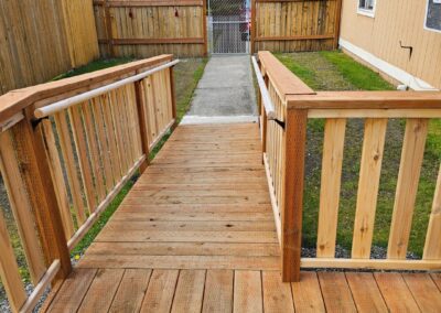 A wooden ramp with railings leads from a house to a fenced backyard with a concrete path and a chain-link gate.