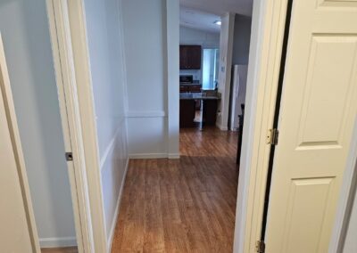 A hallway with wood flooring leads to a kitchen with dark cabinets and stainless steel appliances; doors are open on both sides of the hallway.