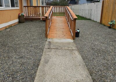 Concrete walkway leading to a wooden wheelchair ramp attached to a tan house, with gravel covering the yard and a white picket fence on the right.