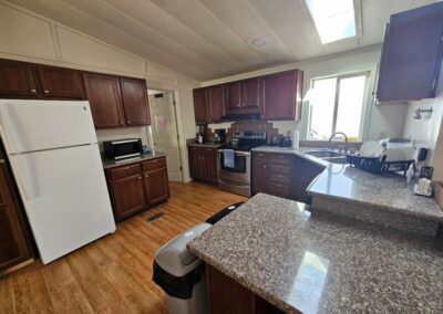 A kitchen with brown cabinets, granite countertops, a white refrigerator, stove, microwave, double sink, and wood flooring, illuminated by natural light from a window and skylight.
