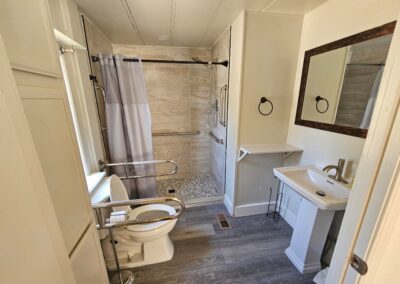 Accessible bathroom with grab bars, raised toilet seat, walk-in shower with a curtain and bench, pedestal sink, wall mirror, and gray wood-style flooring.
