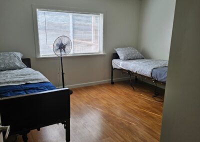 A simple room with two single beds, a standing fan near the window, wood flooring, and white walls.