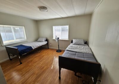 A simple room with two single beds, a standing fan between them, wood-look flooring, and two windows allowing natural light.